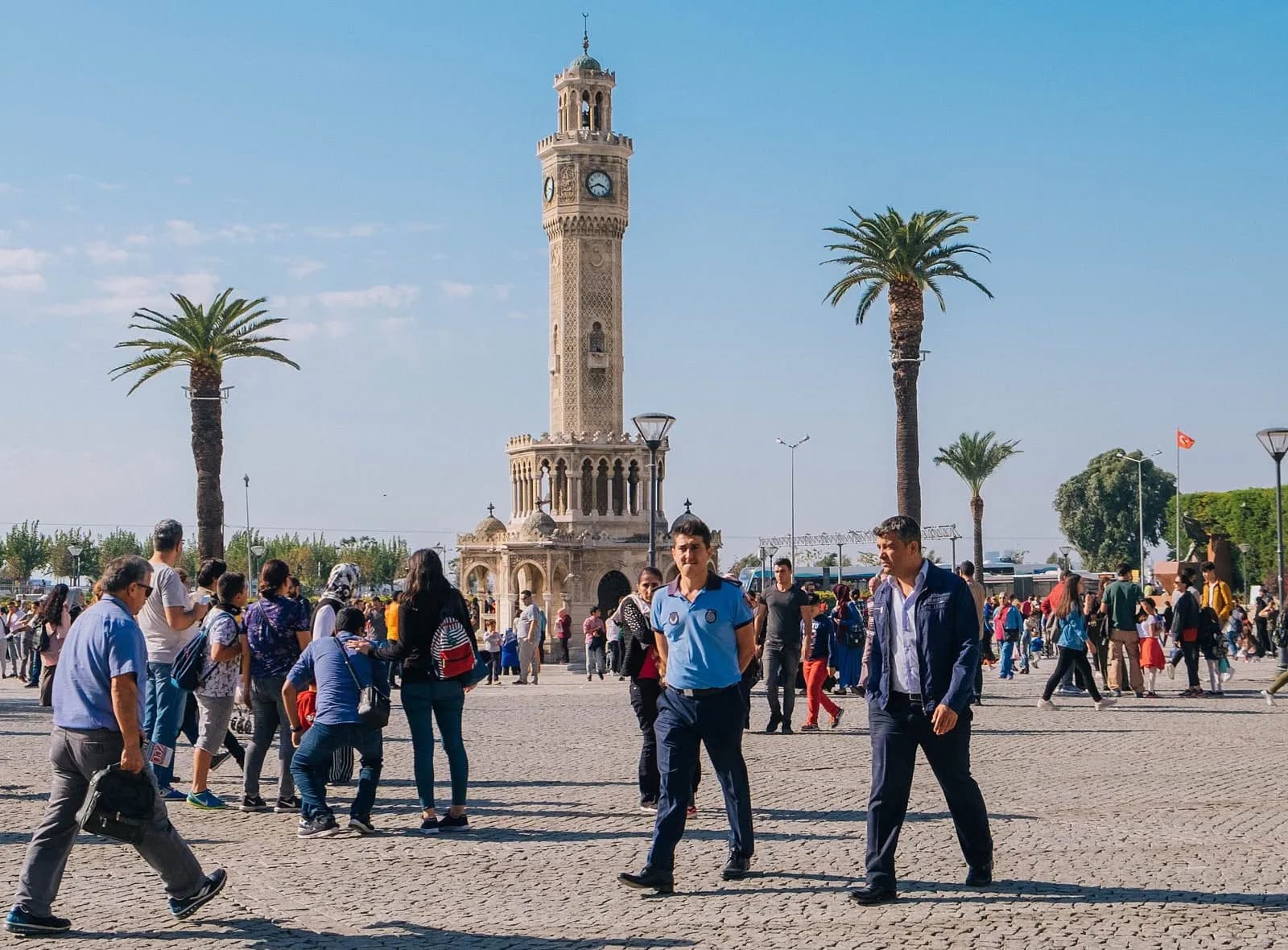 Der Uhrenturm von Izmir (İzmir Saat Kulesi) überragt den Konak-Platz, umgeben von Palmen und Tauben, mit dem Izmir-Golf im Hintergrund